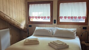 Un dormitorio de cabaña de madera con una cama grande, dos ventanas con cortinas estampadas de corazones y un radiador.