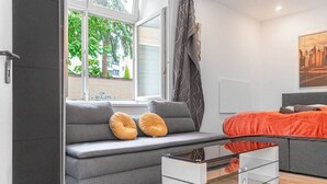Ein modernes Zimmer mit grauem Sofa, einem Glastisch, einem Bett mit orangefarbener Bettwäsche und einem Fenster mit Blick auf Bäume und Gebäude.