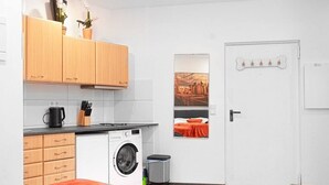 Ein kleines Zimmer mit Waschmaschine, einem Bett mit orangefarbener Decke, einer kleinen Küchenzeile und einer Tür mit knochenförmiger Dekoration.