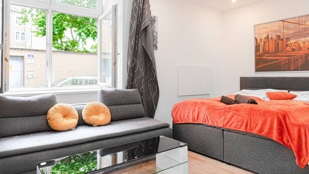 Ein modernes Schlafzimmer mit grauem Sofa, einem Glastisch, einem Bett mit orangefarbener Decke und einem Stadtbildgemälde.