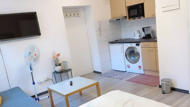 A compact living space with a sofa, a small table, a washing machine, a microwave, and a TV mounted on the wall.