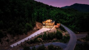 A modern two-story house with a large glass facade, surrounded by a forested landscape and a winding road. - Cozy Luxe Home,Breathtaking Mtn. Views of fall foliage, Outdoor sauna,large deck (Sevierville)
