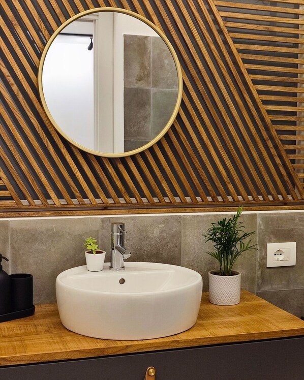Un baño con un espejo redondo, un lavamanos blanco, una planta en maceta y un techo de listones de madera.