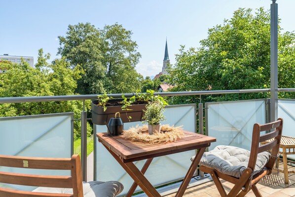 A balcony with a wooden table and chairs, a potted plant, and a view of a church spire.