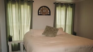 Quarto com cama grande, cortinas verdes, uma pequena mesa com planta e decoração na parede.