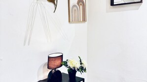 Eine weiße Wand mit einem gerahmten abstrakten Kunstwerk, einer Pendelleuchte, einer Lampe und einer kleinen Vase mit Blumen.