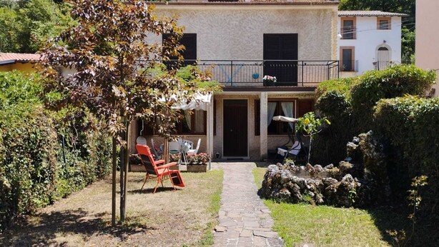 Une cour avec un chemin de pierre menant à un bâtiment doté d’un balcon. On y trouve des chaises et une table à l’extérieur, sous un arbre.