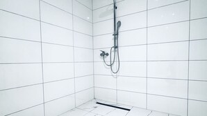 A bathroom with white tiled walls and floor, a shower with a handheld showerhead, and a drain in the center.