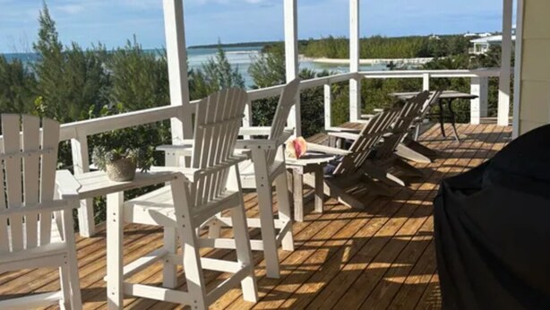 Una terraza de madera con sillas Adirondack blancas y una mesa, con vistas a una playa y al océano.