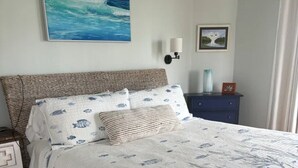 Un dormitorio con cama, mesita de noche, cómoda, un cuadro del océano y una lámpara de pared.