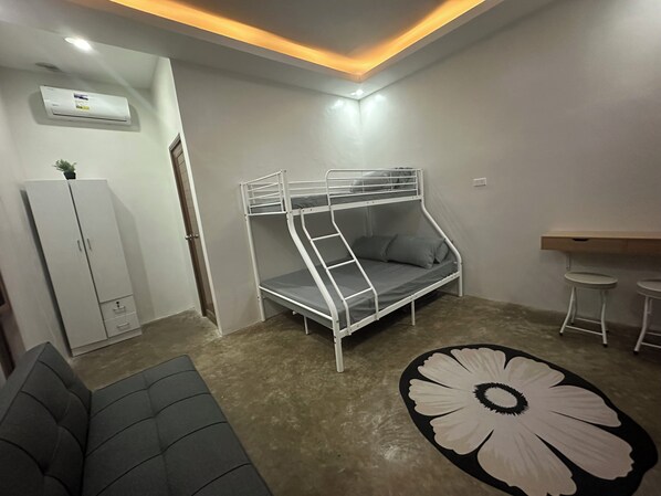 A bunk bed with a desk and two stools, a sofa, and a flower-patterned rug in a room with white walls and a ceiling light.