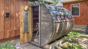 Una estructura con forma de barco de madera, con una puerta, rodeada de un jardín con valla de madera y una casa al fondo.