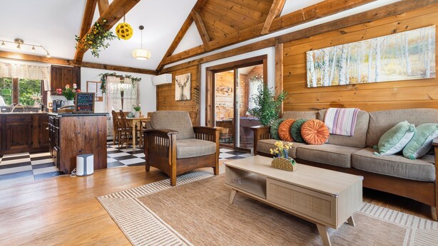 A cozy cabin interior with wooden beams, a dining area, a sofa with cushions, and a coffee table.