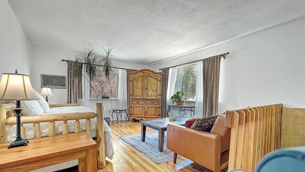 Une pièce chaleureuse avec un lit en bois, un banc en bois, un canapé en cuir et une petite table. On y trouve une fenêtre avec des rideaux, une plante en pot et un radiateur.