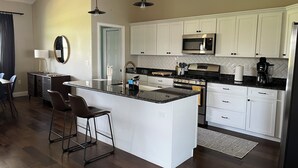 A modern kitchen with white cabinets, a central island, stainless steel appliances, and a dining area with bar stools. - Sunny Daze lake time is always a great time! (Lake Placid)