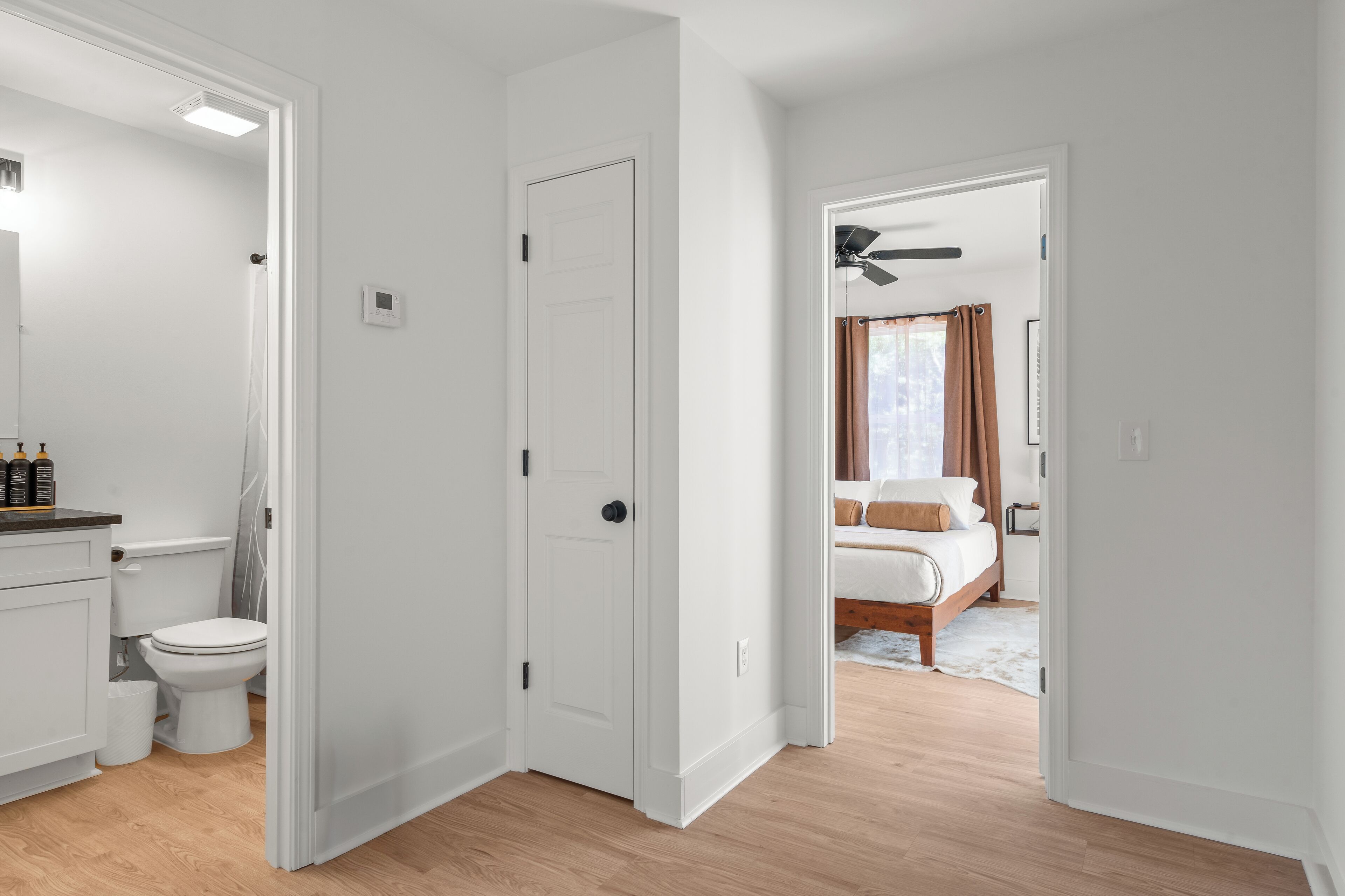 A hallway leading to a bedroom with a bed, a ceiling fan, and a bathroom with a toilet and sink.