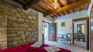 A room with a stone wall, wooden ceiling beams, a bed with a red blanket, and a dining area visible through an open door. - Appartamento rustico con piscina nell'antico "Podere la Querciola, Guardistallo" (Guardistallo)