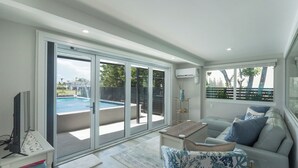 A modern living room with a pool view, a flat-screen TV, and a sofa with cushions. - ☀️Private Pool | Pet Friendly | Curlew Beach Cottage☀️ (Fort Myers Beach)