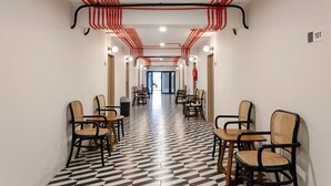 Un pasillo con piso a cuadros, sillas y mesas, con tuberías rojas y negras en el techo.