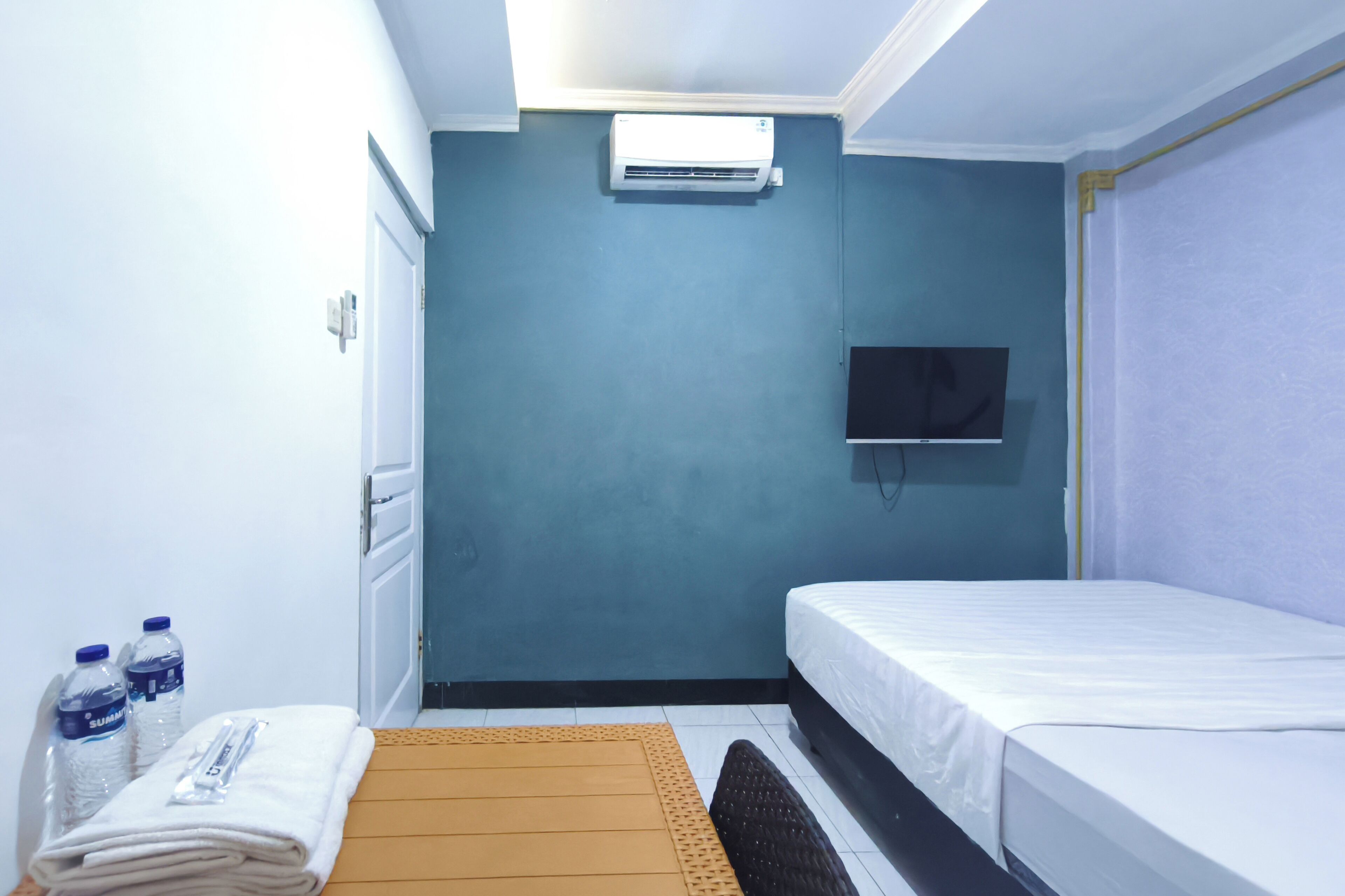 A hotel room with a bed, a flat-screen TV mounted on the wall, an air conditioning unit, a window, and a small table with water bottles and a towel.