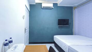 A hotel room with a bed, a flat-screen TV mounted on the wall, an air conditioning unit, a window, and a small table with water bottles and a towel.
