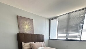 Ein Schlafzimmer mit einem Bett, einem kleinen Tisch, einer Deckenleuchte, einem Fenster mit Jalousien und einem Wandbild.