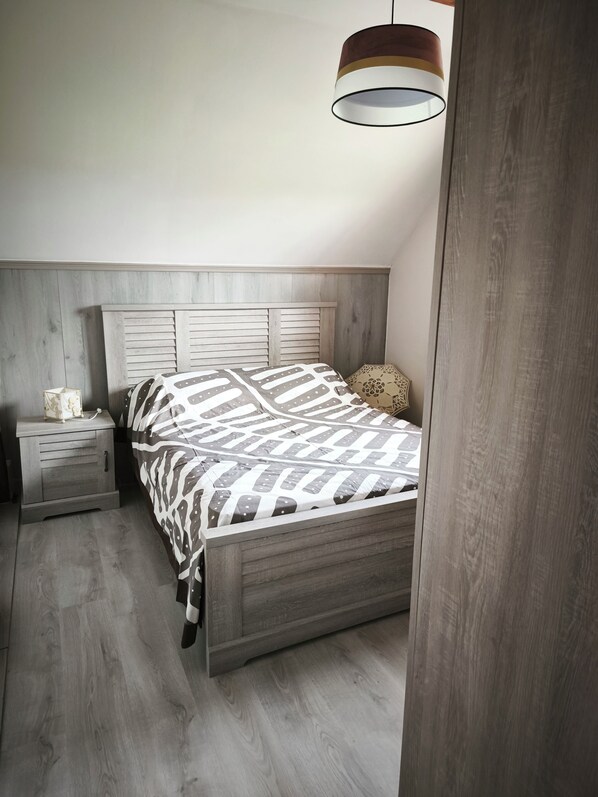 Een slaapkamer met een houten hoofdbord, een bed met beddengoed met een patroon, een nachtkastje met een lamp en een houten vloer.