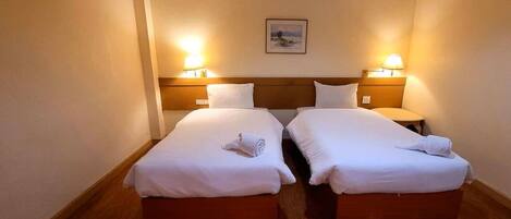 A hotel room with two beds, each with a wooden headboard, white linens, and a pair of bedside lamps.