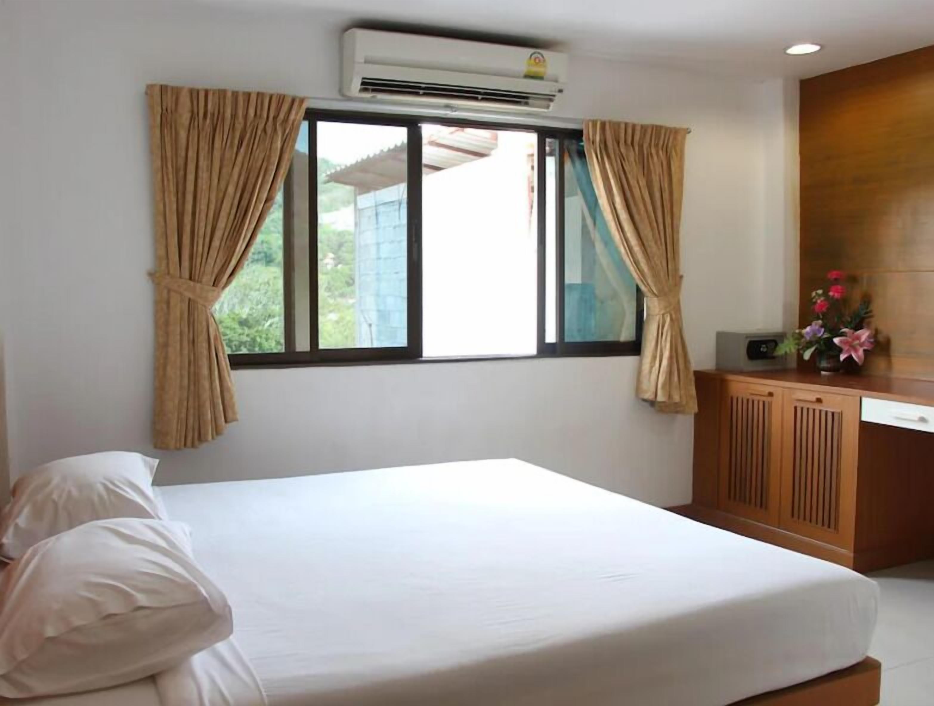 Une chambre d’hôtel avec un lit, des rideaux, un appareil de climatisation, une fenêtre et une petite armoire sur laquelle est posé un vase de fleurs.