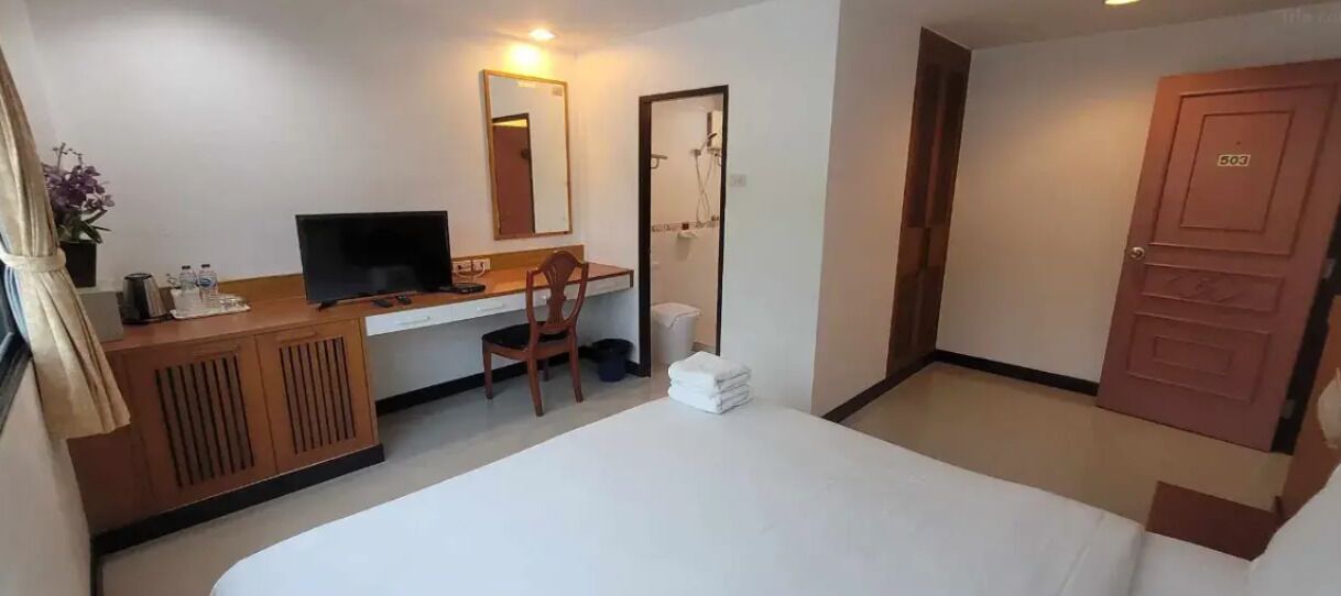 A hotel room with a bed, a desk with a chair, a television, a mirror, and a shower area.
