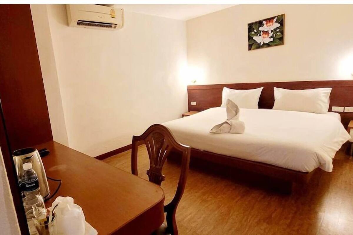 A hotel room with a bed, bedside table, chair, and an air conditioning unit.