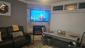 Ein Wohnzimmer mit Kamin, einem an der Wand befestigten Fernseher, einem Esstisch mit Stühlen und einer Couch mit Überwurfdecke.