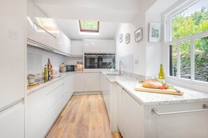 A modern kitchen with white cabinetry, a wooden floor, a large window with a view of greenery, and a countertop with a loaf of bread and a bottle of olive oil.