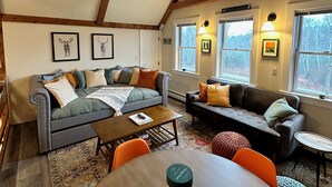 A cozy loft-style living room with exposed wooden beams, a gray sofa, a coffee table, and a view of a forest through large windows. - Three Peaks Hideaway, a Tranquil 4-bedroom house with Mountain Views in Peru (Peru)