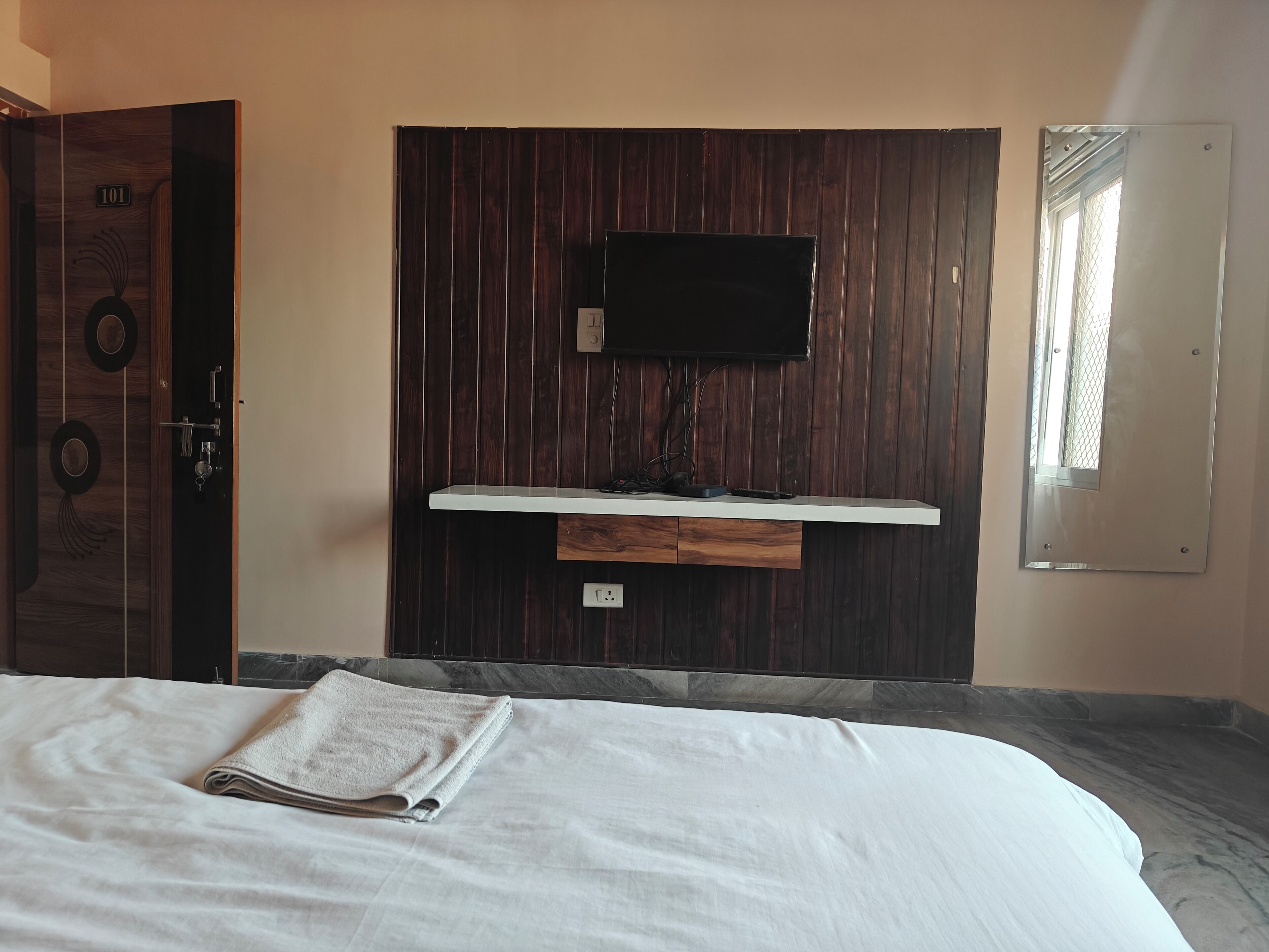 A bedroom with a bed, a flat-screen TV mounted on a wooden panel wall, a window with a mesh screen, and a door with a number sign.
