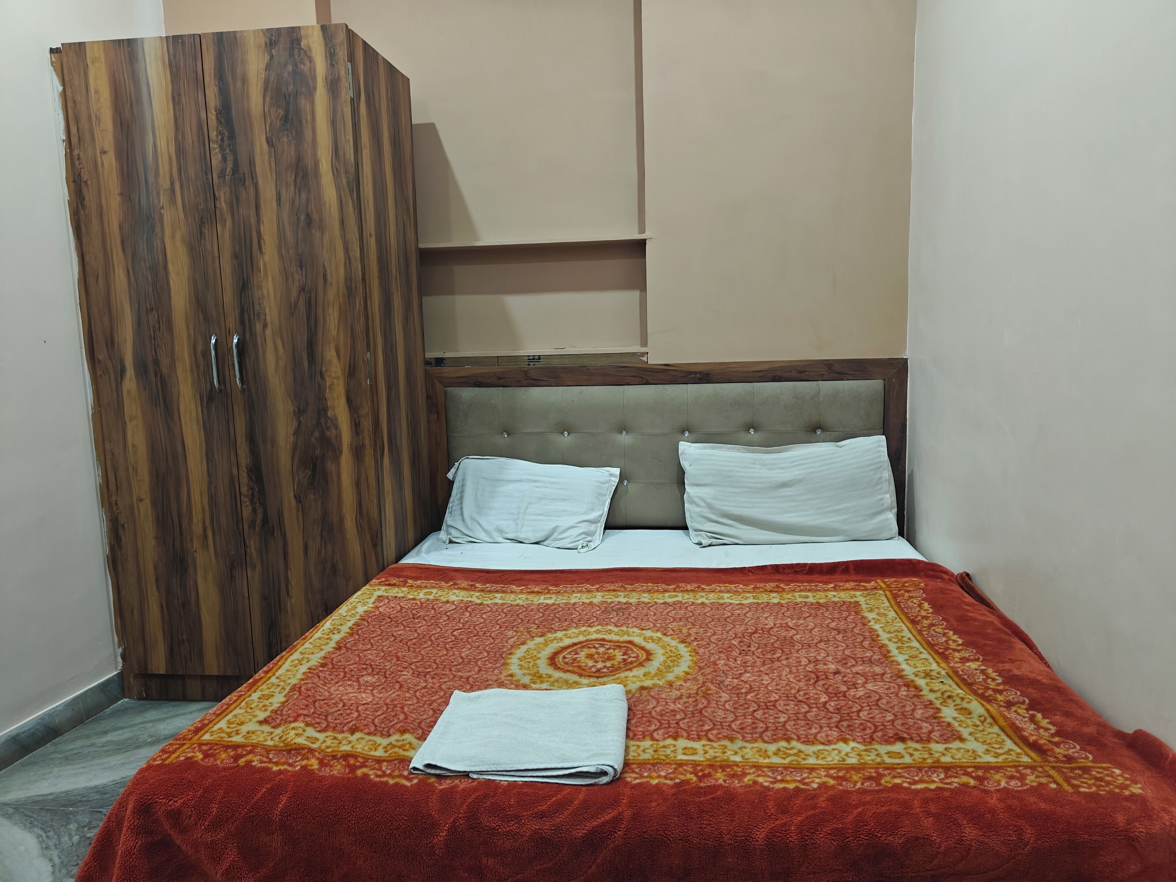 Una camera da letto con un letto, due cuscini, una coperta a letto con motivi rossi e gialli, un asciugamano piegato e un armadio in legno.