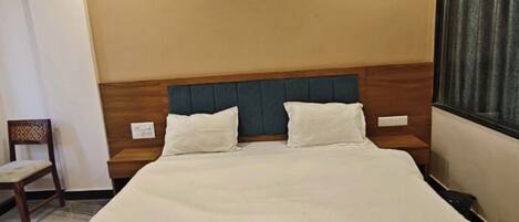 A hotel room with a large bed, a wooden headboard, an air conditioning unit, a chair, and a window with curtains.
