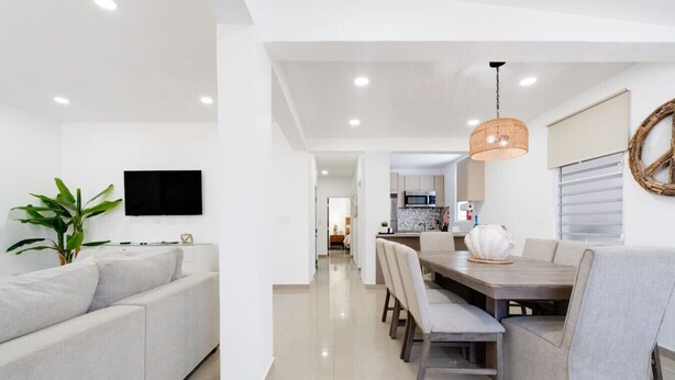 Una sala de estar moderna con un sofá, una mesa de comedor con sillas, un televisor montado en la pared y una cocina al fondo.