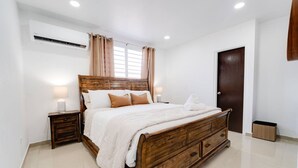 Un dormitorio con cama de madera, paredes blancas, una ventana con cortinas, una puerta y un equipo de aire acondicionado.