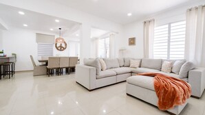 Amplia sala de estar con un sofá modular, comedor con mesa y sillas, y cocina al fondo.