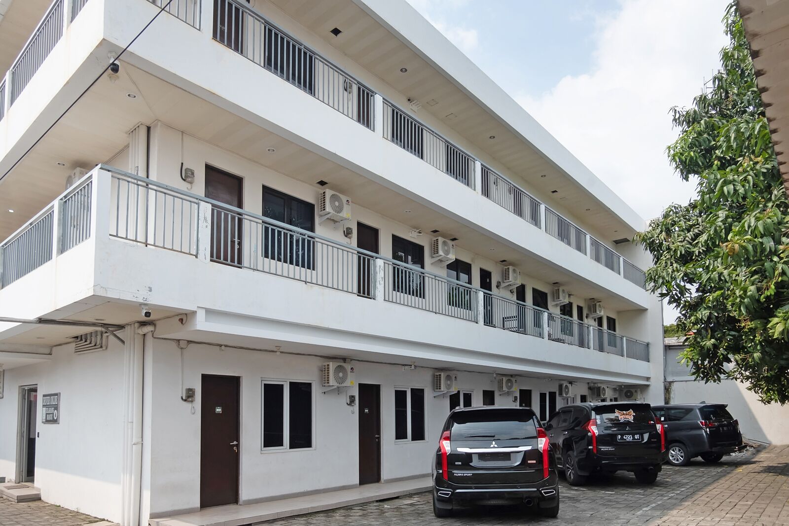 A multi-story building with balconies, air conditioning units, and a parking area with cars.