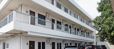 A multi-story building with balconies, air conditioning units, and a parking area with cars.