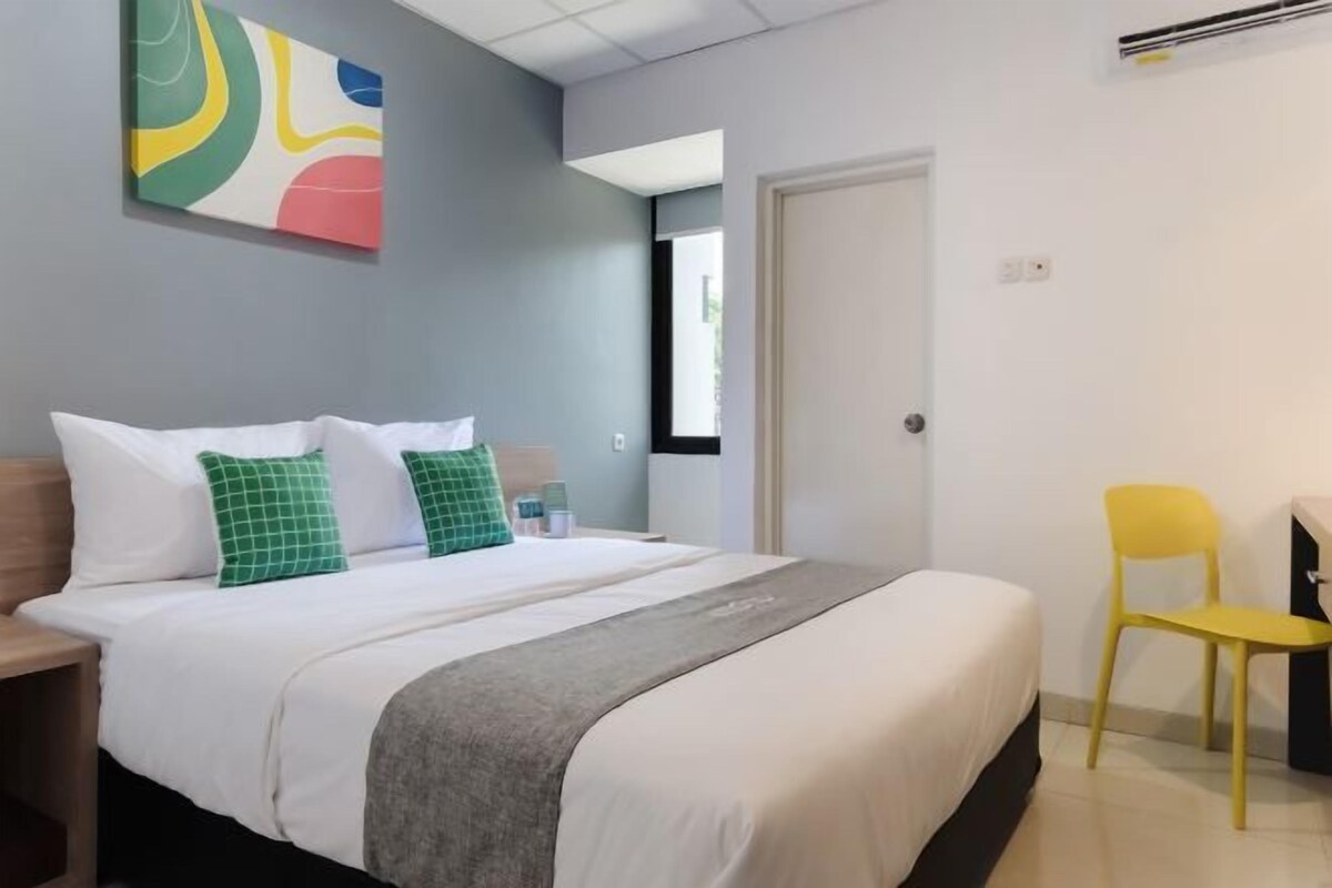 A hotel room with a bed, a desk, a chair, a colorful abstract painting, and a door.