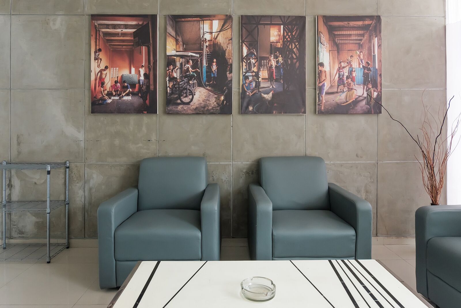 A modern lobby with two grey armchairs, a white table with a glass ashtray, and three large framed artworks depicting indoor scenes.
