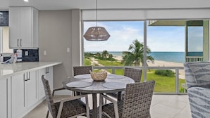 Una cocina moderna con armarios blancos, un comedor con sillas de mimbre y un amplio ventanal con vistas a la playa.
