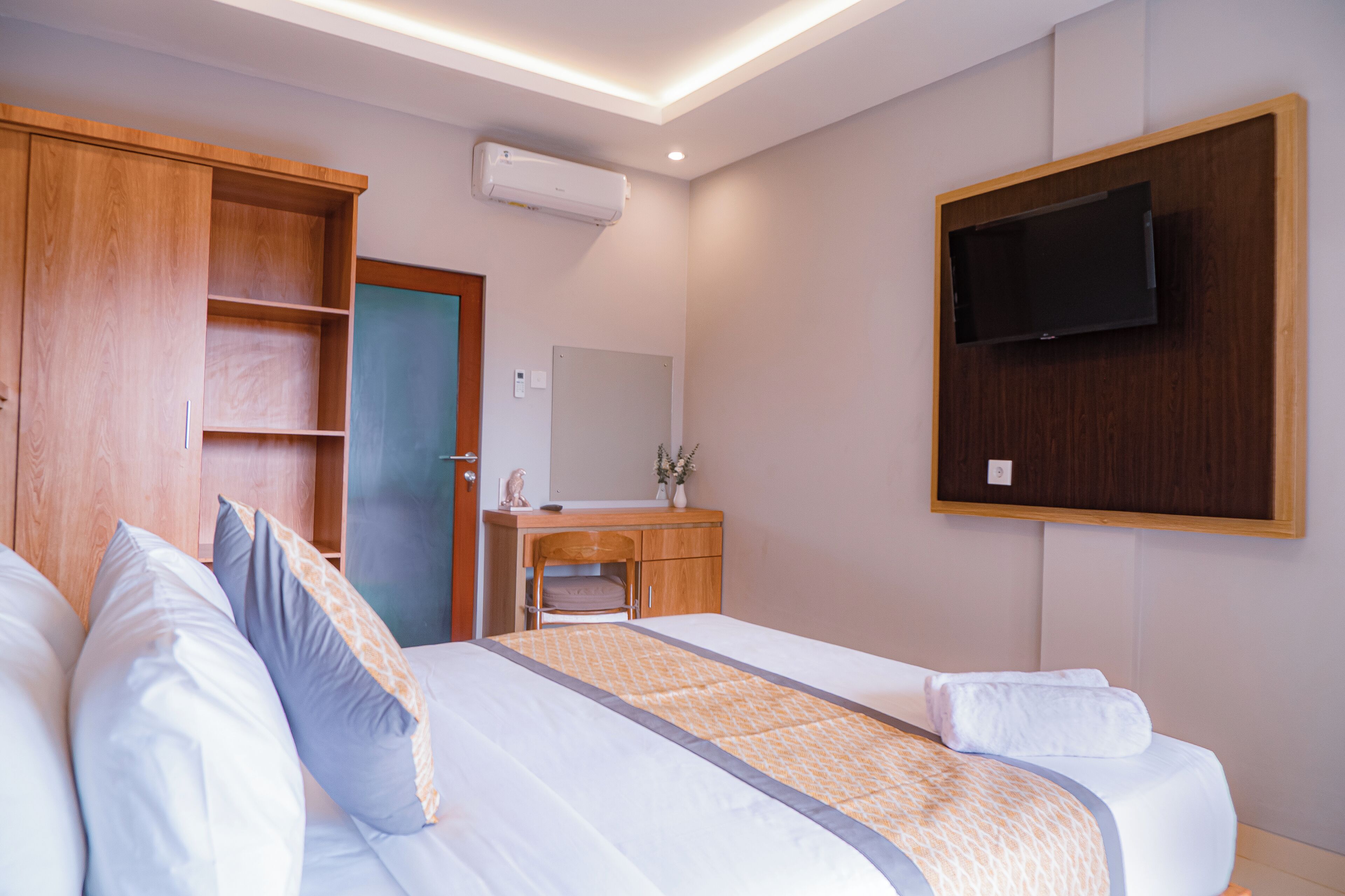 Quarto de hotel com cama, guarda-roupa de madeira, secretária, espelho e televisão presa à parede.