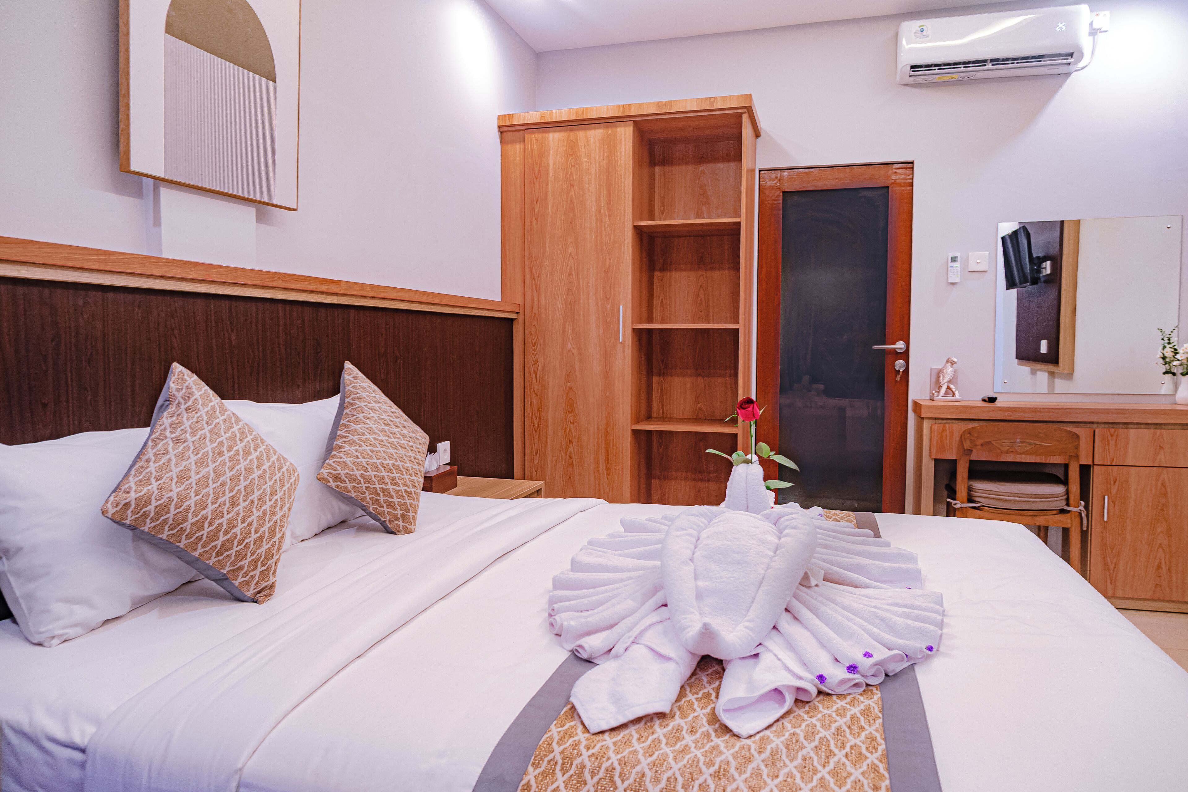 A hotel room with a neatly made bed, a wooden wardrobe, a vanity with a mirror, and an air conditioning unit.