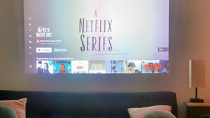 Una sala de estar con un televisor grande mostrando la interfaz de una serie de Netflix, un sofá con cojines, una mesa de centro con un altavoz y algunos objetos encima, y una lámpara sobre una mesita auxiliar.