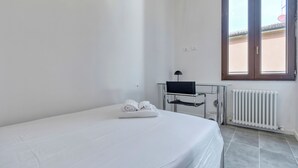 A hotel room with a bed, a desk with a computer, a window, and a radiator. - Suite Clapton | BluesHouse Carracci Suites (Bologna)