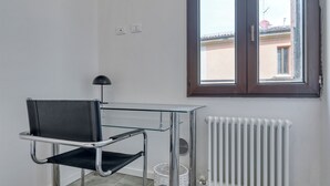 A room with a black leather chair, a glass table, a window with a view of buildings, and a white radiator. - Suite Clapton | BluesHouse Carracci Suites (Bologna)